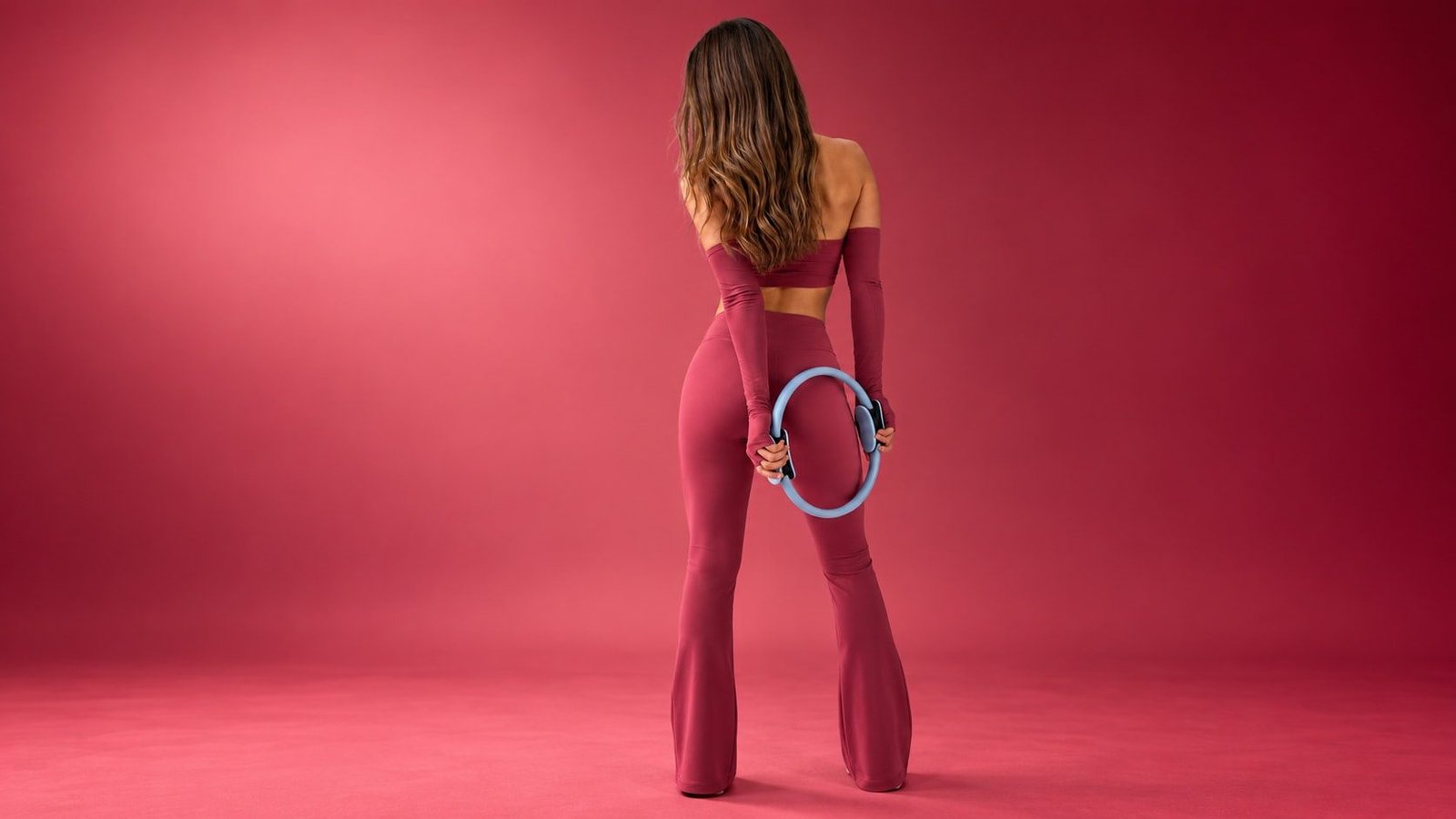 Eine Frau in Bordeaux mit langem Haar steht mit dem Rücken zur Kamera und hält einen blauen Pilates-Ring hinter dem Rücken.